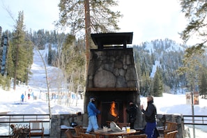 Une cheminée en pierre avec un feu, entourée de chaises et de tables en bois, le tout se dressant sur fond de montagnes enneigées.