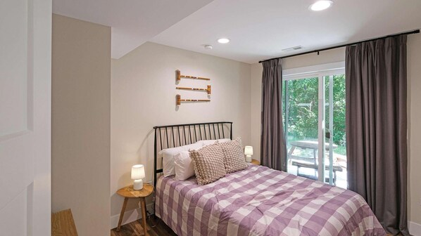 A bedroom with a bed, a nightstand, a lamp, a window with curtains, and a view of greenery outside.