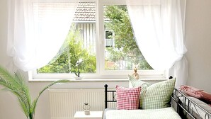 Une chambre à coucher avec un lit, une table de chevet, un radiateur, une fenêtre avec des rideaux et une plante en pot.