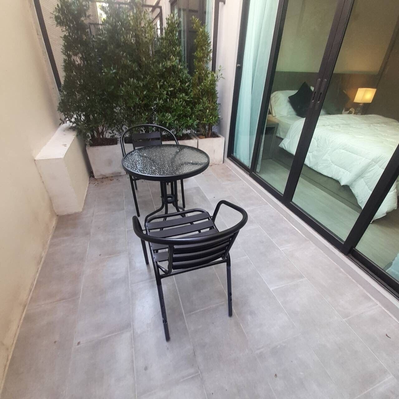 Un balcone con un tavolo rotondo e due sedie, una pianta in vaso, e un letto visibile attraverso una porta in vetro.