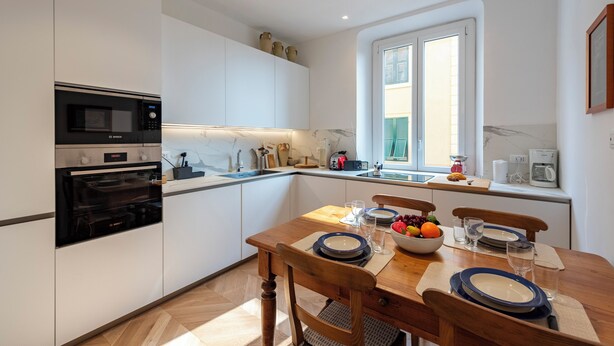 Une cuisine moderne avec des meubles blancs, une table à manger en bois mise avec des assiettes, des bols et des verres, et une fenêtre avec des stores.