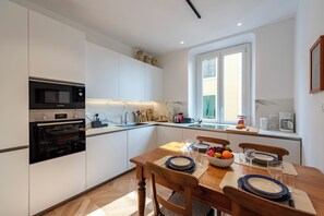 A modern kitchen with white cabinetry, a wooden dining table set with plates, bowls, and glasses, and a window with blinds. - Castelletto House Genova City Center (Genoa)