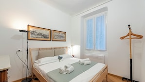 Un lit bien fait, avec du linge de lit blanc et un plaid vert, un porte-manteau en bois et deux tableaux encadrés accrochés au mur.