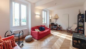 Un salon spacieux avec un canapé rouge, un parquet en bois disposé en chevrons, un bureau avec une lampe, une bibliothèque et une vue dégagée grâce à de grandes fenêtres.