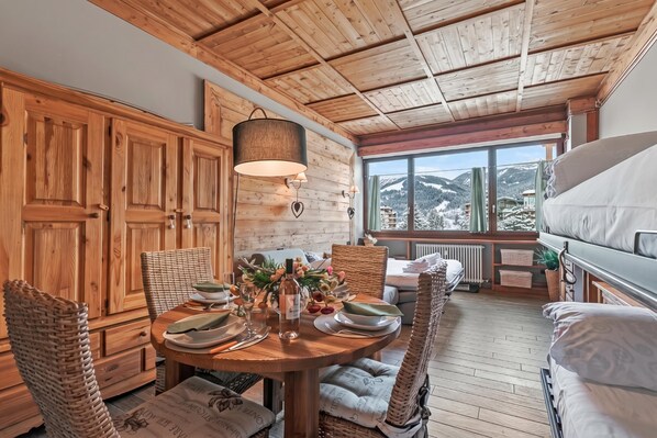 A cozy dining area with a wooden table set for four, surrounded by wicker chairs, a hanging light fixture, and a view of snowy mountains through large windows.