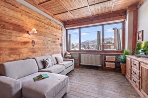 A cozy room with a wooden ceiling, a sofa, a window with a view of snow-covered mountains, and a radiator.