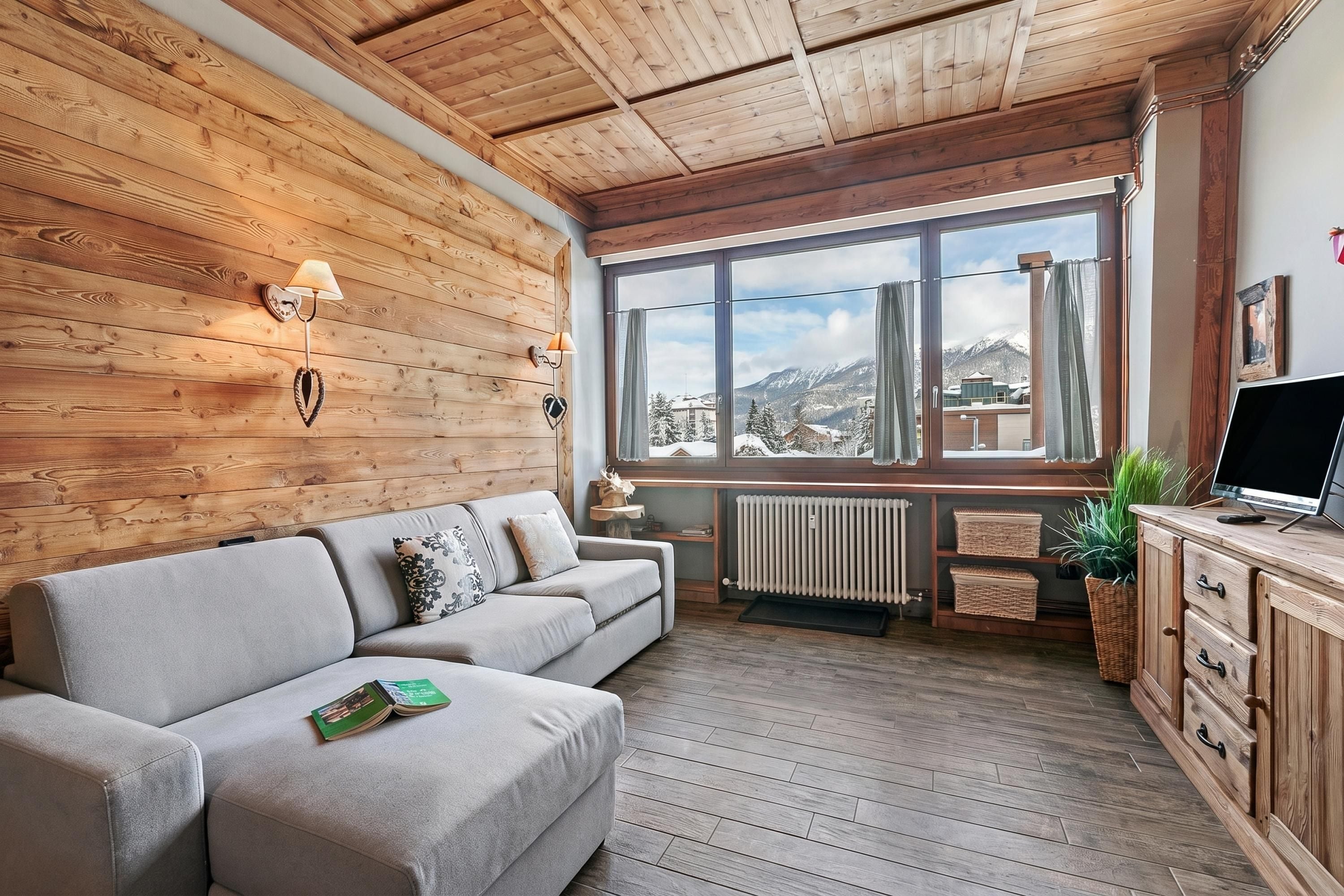 Ein gemütliches Zimmer mit Holzboden, einem grauen Sofa, einem Fenster mit Blick auf schneebedeckte Berge und einer Holzkabine mit Fernseher.