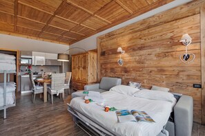 A hotel room with a wooden ceiling, a bed with white bedding, a grey sofa, a dining table with chairs, and a kitchenette. - Studio Charming Sportinia Town Center (Sauze d'Oulx)