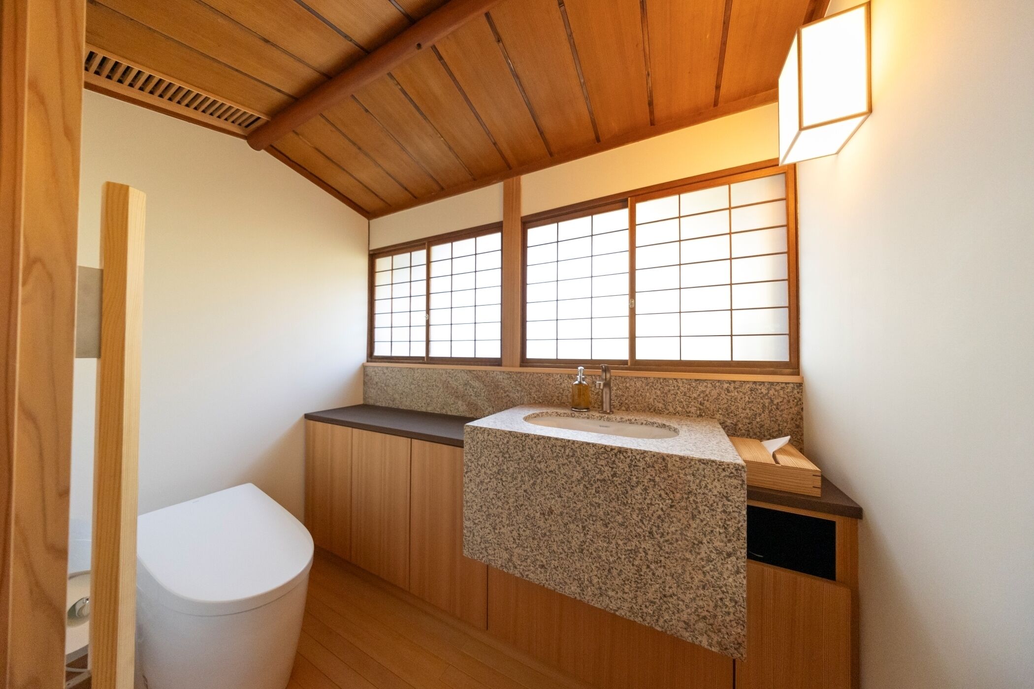 A compact bathroom with a wooden ceiling, a white toilet, a granite countertop sink, and a window with a grid pattern.