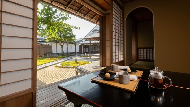 A traditional Japanese room with tatami flooring, a low table with a tea set, and a view of a garden and traditional buildings. - mameya (Sasayama)