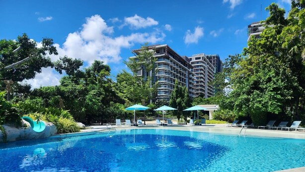 Una piscina azul cristalina rodeada de abundante vegetación y un edificio moderno con varios balcones.