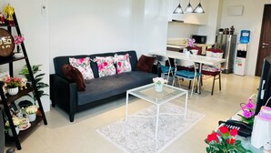 Una sala moderna con un sofá gris oscuro, una mesa de centro de vidrio y un tapete blanco. Hay una zona de comedor con una mesa y sillas blancas, y una cocina con un refrigerador y dispensador de agua.