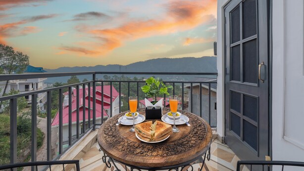 A balcony with a table set for two, a sandwich, and two glasses of orange juice, overlooking a mountainous landscape. - The Chalet Homestay (Kasauli)