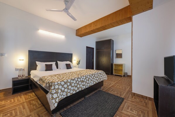 Una habitación de hotel moderna con una cama grande, suelo de madera, ventilador de techo, televisor y armario.