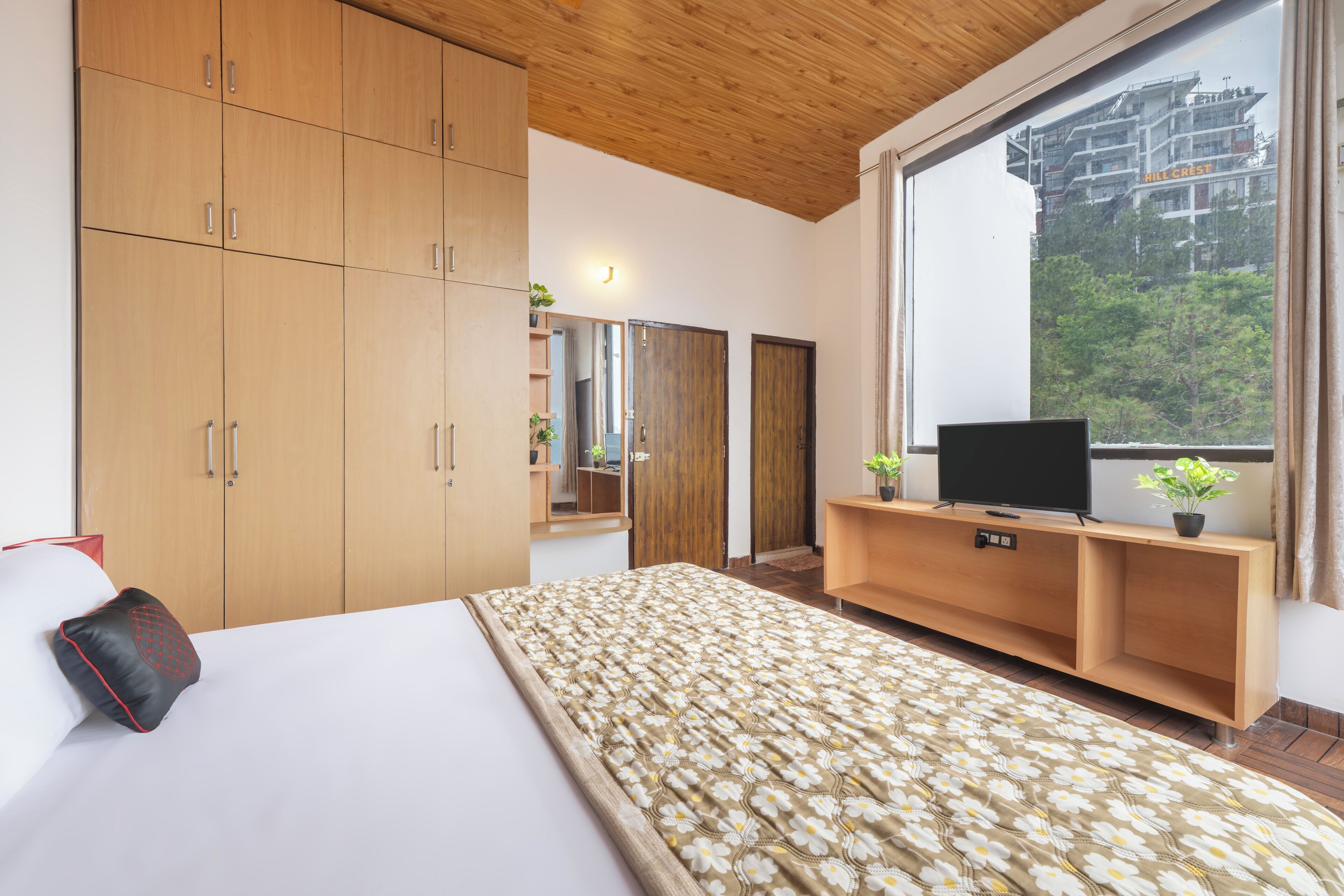 A hotel room with a large bed, wooden wardrobe, a TV on a wooden console, and a large window with a view of buildings.