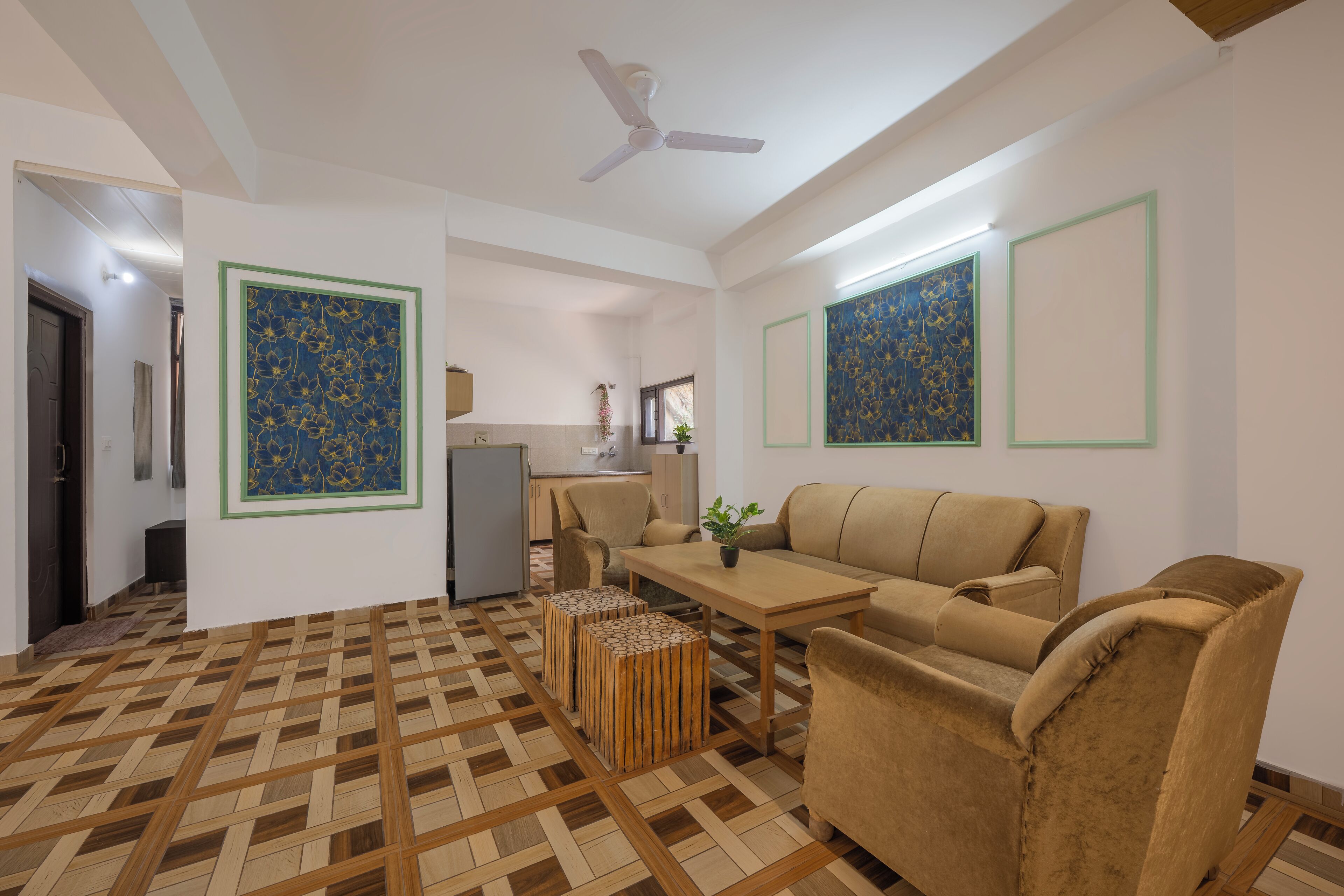 Un salon avec un canapé, un pouf et une table. On y trouve un ventilateur de plafond, une cuisine à l’arrière-plan et un sol à motifs.