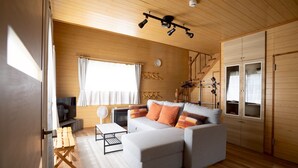 A modern living room with a sectional sofa, wooden flooring, a staircase, and a large window with curtains. - LAKE TOYA PREMIUM CABIN (Toyako)