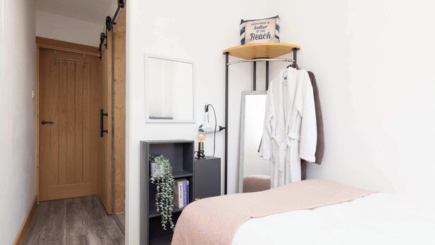 A bedroom with a wooden door, a bedside table with a lamp, a mirror, and a shelf with books and a plant. - Coastal Retreat with Hot Tub, Walk to Beach (West Wittering)