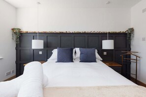 Un lit bien fait, avec une literie blanche et deux oreillers bleus, une tête de lit en bois foncé ornée d’une guirlande décorative, et une table de chevet en bois avec une lampe suspendue.