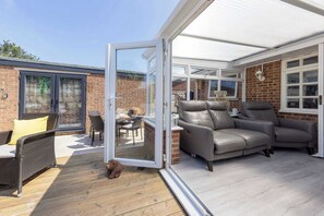 Une terrasse couverte avec une porte vitrée donnant sur une façade en briques, des canapés en cuir gris et un fauteuil en osier.