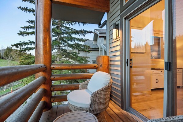 A wooden deck with a wicker chair and a small table, a glass door leading to a kitchen, and a view of trees and a house in the background.