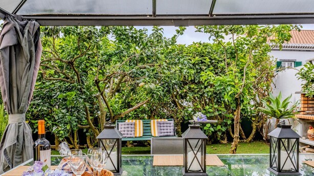 An outdoor dining area with a glass table, two cushioned benches, and a bottle of wine.