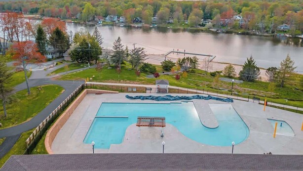 Ein Schwimmbecken mit Rettungsschwimmerstand, umgeben von einer Rasenfläche und Blick auf einen See mit Häusern im Hintergrund.