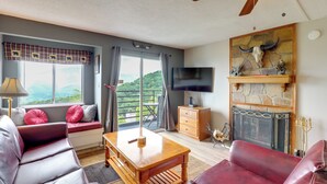 Ein Wohnzimmer mit einer Couch, einem Holztisch, einem Kamin mit Steinverkleidung, einem Fernseher und Blick ins Grüne.