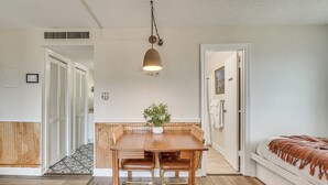Une pièce avec une table à manger en bois, deux chaises, une plante en pot, un lit avec une couverture et une porte blanche.