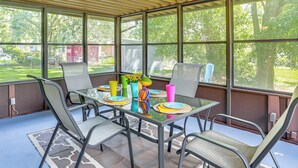 Un patio cubierto con una mesa de vidrio y sillas de metal, rodeado de amplias ventanas que dan a un jardín.