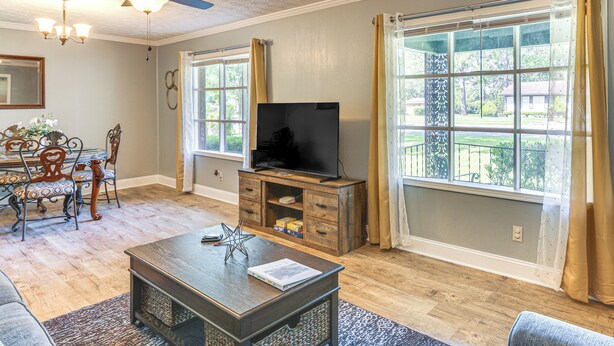 Una sala de estar con televisor sobre un mueble de madera, un comedor con mesa y sillas, y ventanas amplias con cortinas.