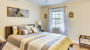Un dormitorio con una cama grande, un ventilador de techo, una ventana con cortinas, una mesita de noche con una lámpara y un cuadro en la pared.