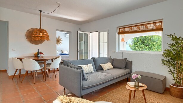 A living room with a grey sofa, a dining table with chairs, a hanging light fixture, a window with a bamboo blind, and a potted plant. - Tres Corazones - Moraira, Costa Blanca (Moraira)
