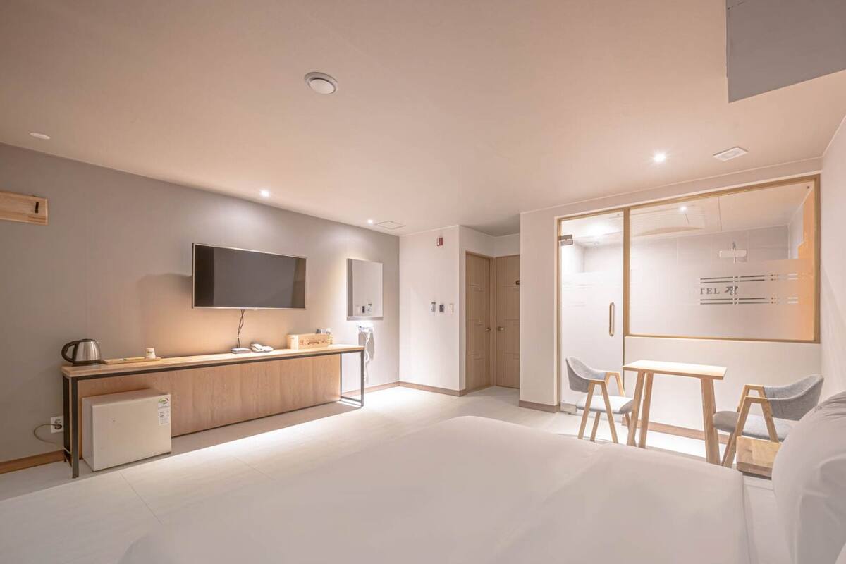 A modern hotel room with a kitchenette, a TV, a desk with chairs, and a large window.