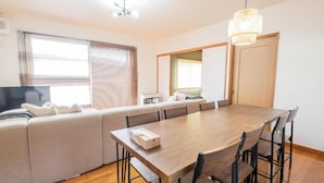 A dining area with a long wooden table and chairs, a sofa with cushions, and a hanging light fixture. - UNO Toyako (Toyako)