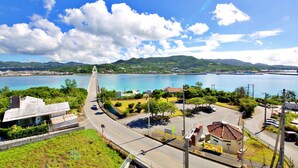 A coastal road with a bridge, a parking area, and a view of the sea and hills. - Ocean Terrace SESOKO 203 (Motobu)
