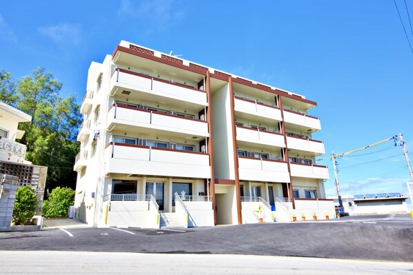 A multi-story hotel with a central staircase, balconies on each floor, and a parking area in front. - Ocean Terrace SESOKO 203 (Motobu)