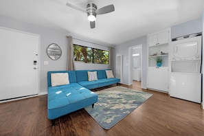 Un espace de vie moderne comprenant un canapé d’angle, un ventilateur de plafond, une grande fenêtre donnant sur un paysage verdoyant et des appareils électroménagers intégrés.