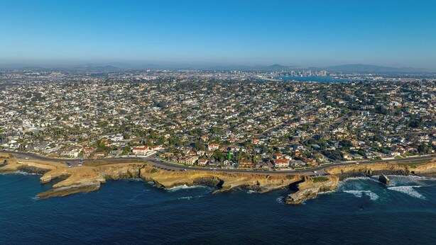 An aerial view of a coastal city with a dense urban area, a prominent coastline with rocky outcrops, and a clear blue sky.