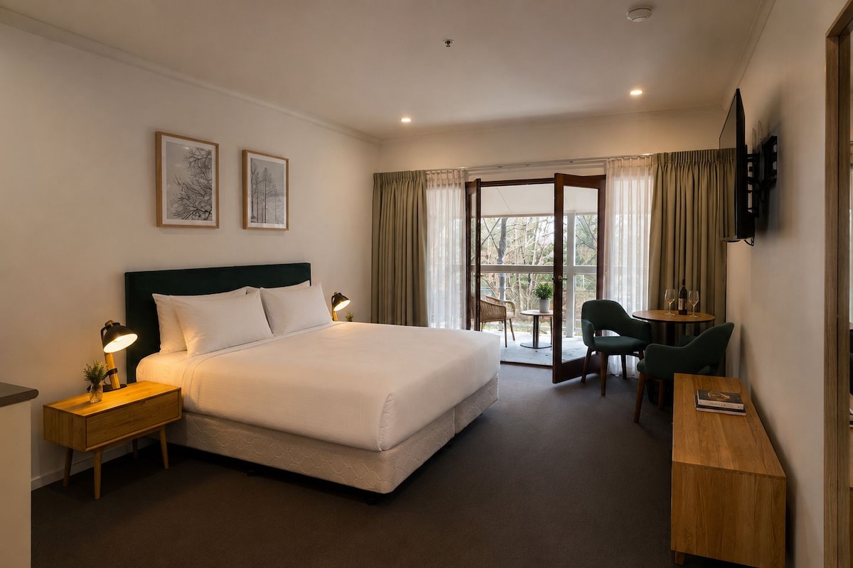A hotel room with a large bed, bedside tables, a desk with two chairs, a TV, and a view through a glass door.