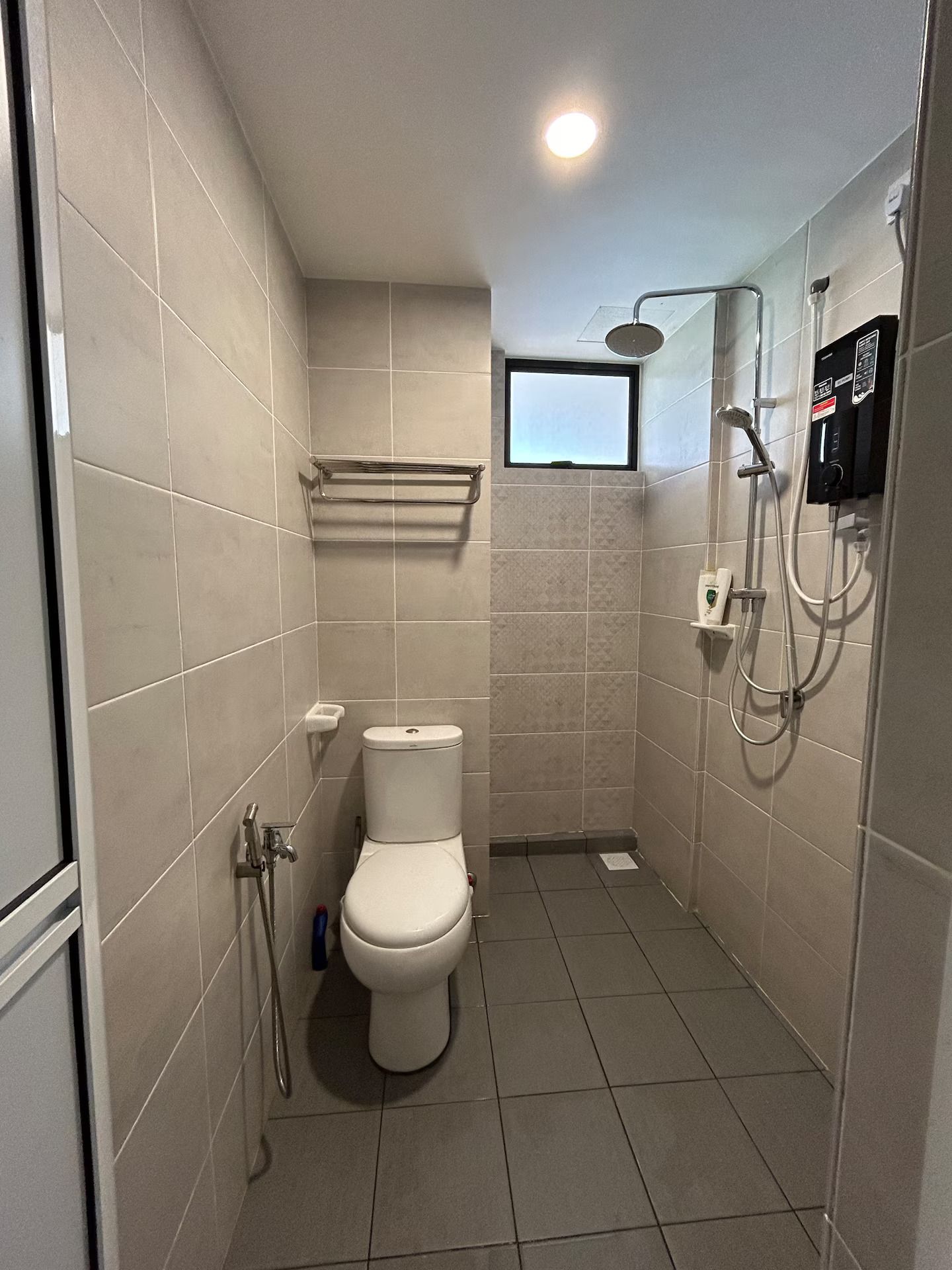 Une petite salle de bain carrelée, équipée de toilettes, d’une douche et d’une fenêtre.