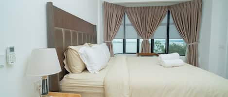 A hotel room with a bed, a bedside table with a lamp, and a view of the outdoors through large windows.