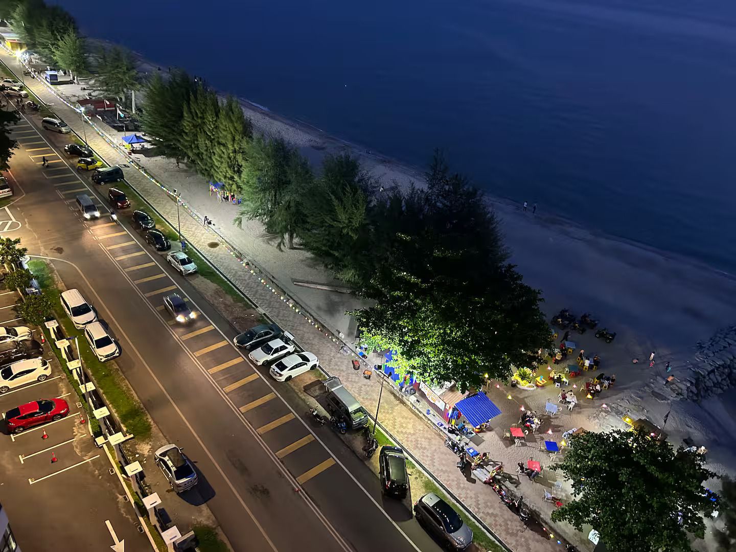 Eine Küstenstraße mit parkenden Autos und einer Strandpromenade, an der sich Menschen und Zelte befinden.