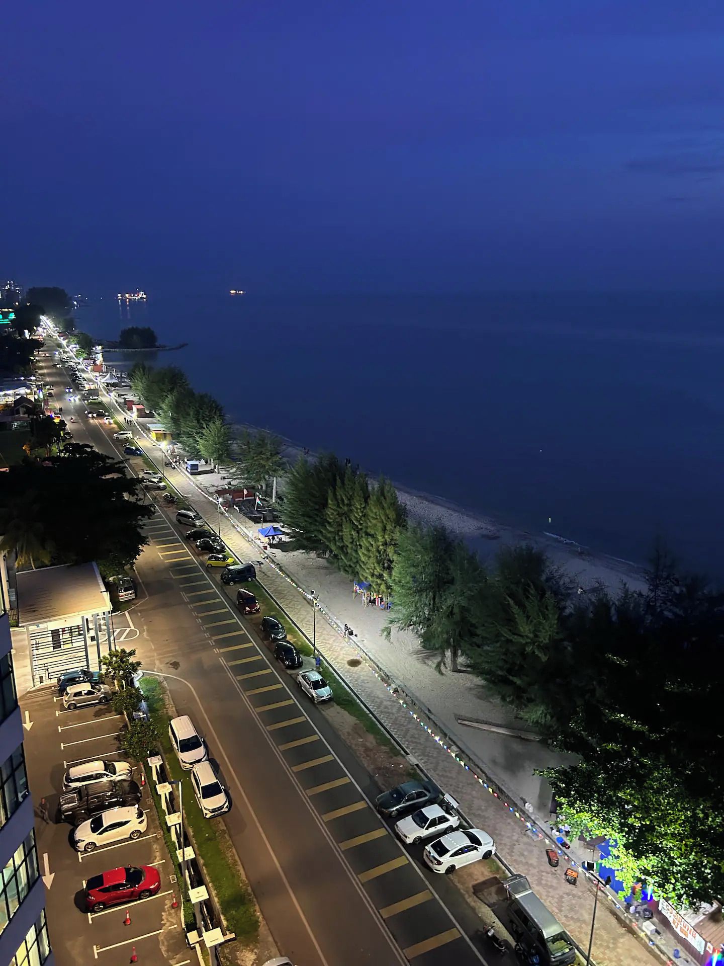 Vue nocturne d’une route côtière, avec des voitures garées et un chemin piétonnier longeant la mer.