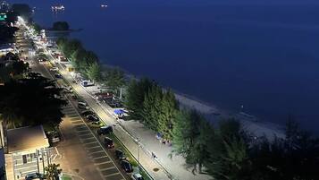 Vue nocturne d’une route côtière, avec des voitures garées et un chemin piétonnier longeant la mer.