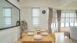 Uma sala de jantar com mesa de madeira posta para quatro pessoas, rodeada por cadeiras, um relógio na parede e uma janela com cortinas.