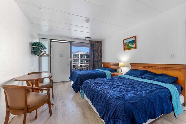 A hotel room with two beds, a wooden chair, a small table, and a view of buildings through a door. - Studio Condo in Prime Location with a Pool (Wildwood)