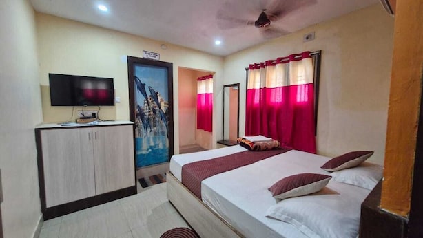 Quarto de hotel com cama, escrivaninha com televisão, quadro na parede e cortinas vermelhas.