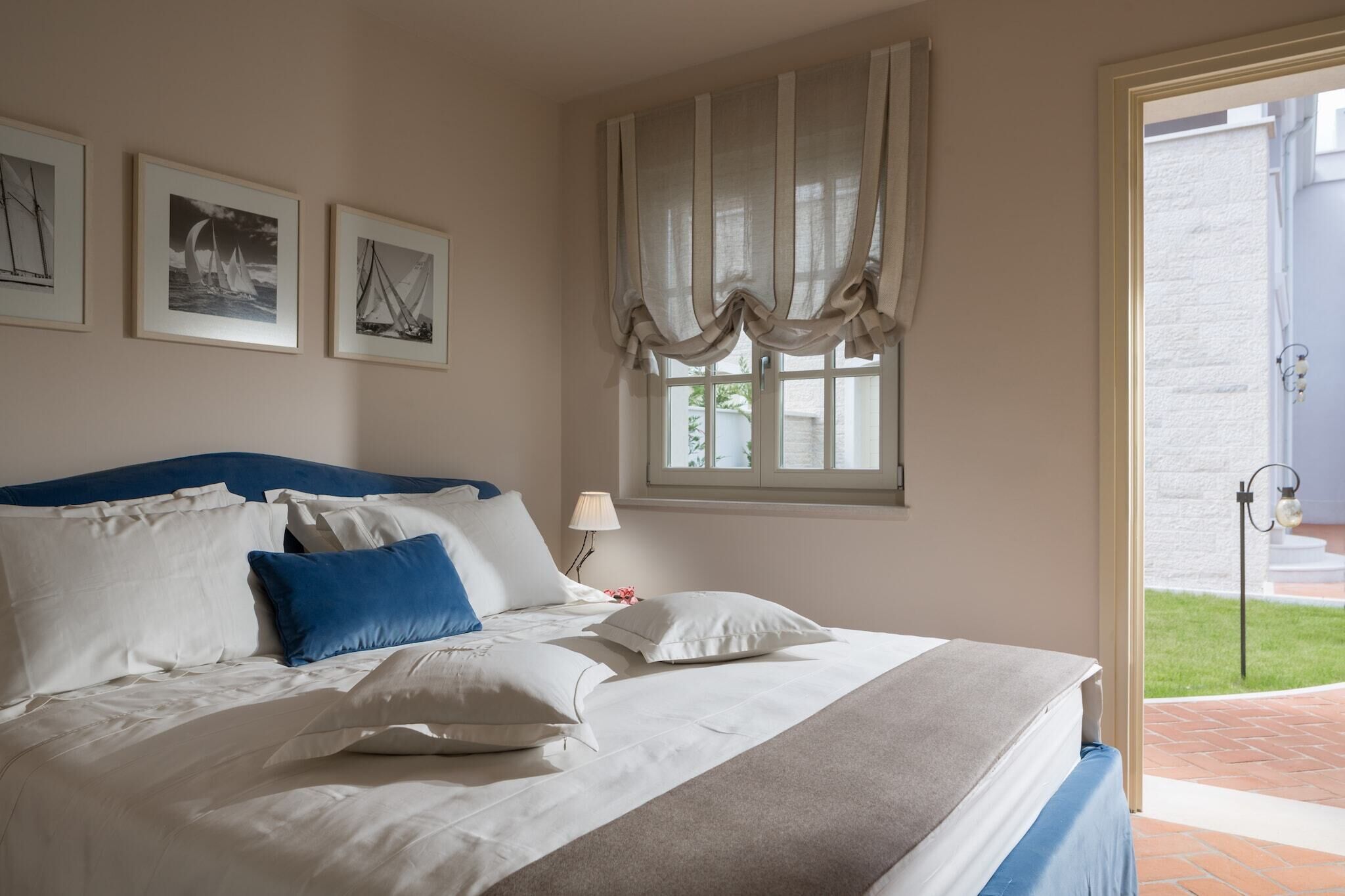 Un letto rifatto con cura, lenzuola bianche e blu, una finestra con tende e tre quadri appesi al muro.