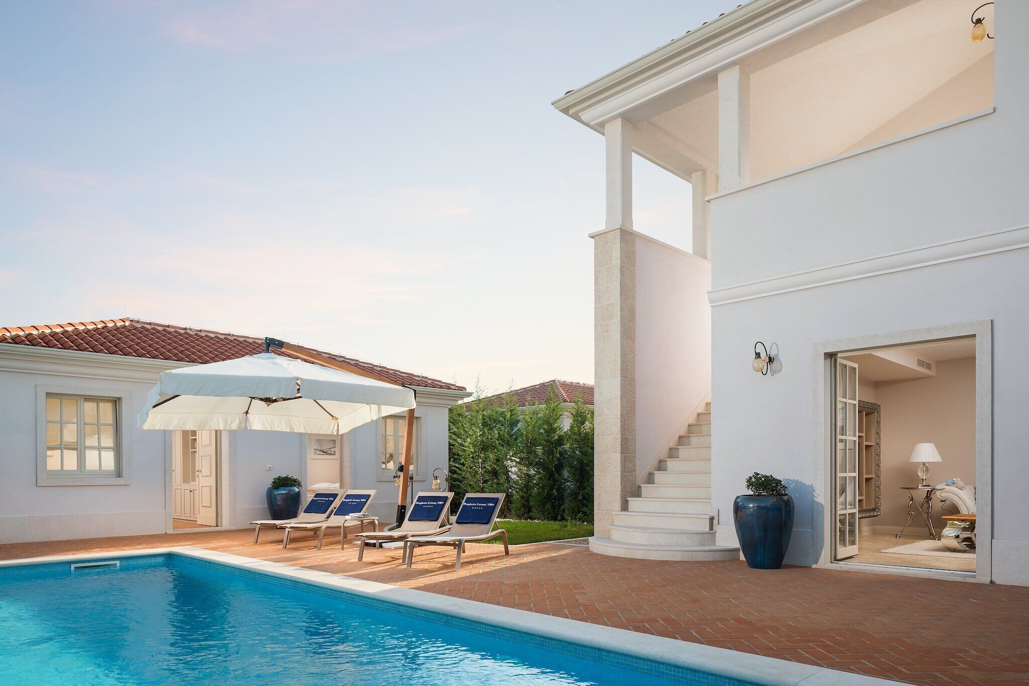 A swimming pool with lounge chairs and umbrellas, a white building with a staircase, and a large potted plant.